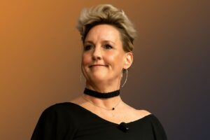 an image of erin brockovich in black outfit wearing a black neck band and brown-black gradient background representing net worth of erin brockovich